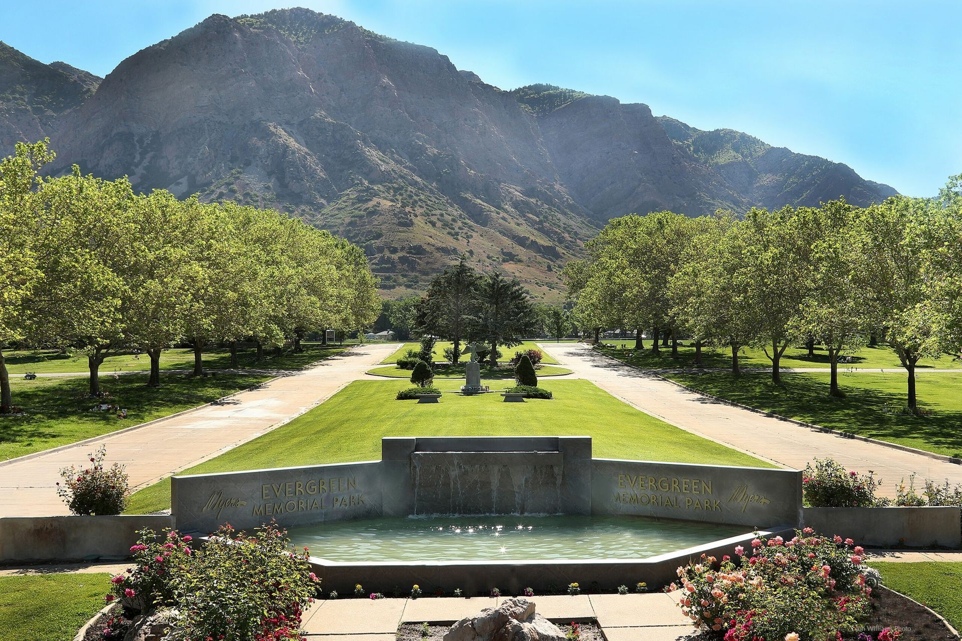 Mountain park with a central fountain, tree-lined paths, and a large rocky peak in the background