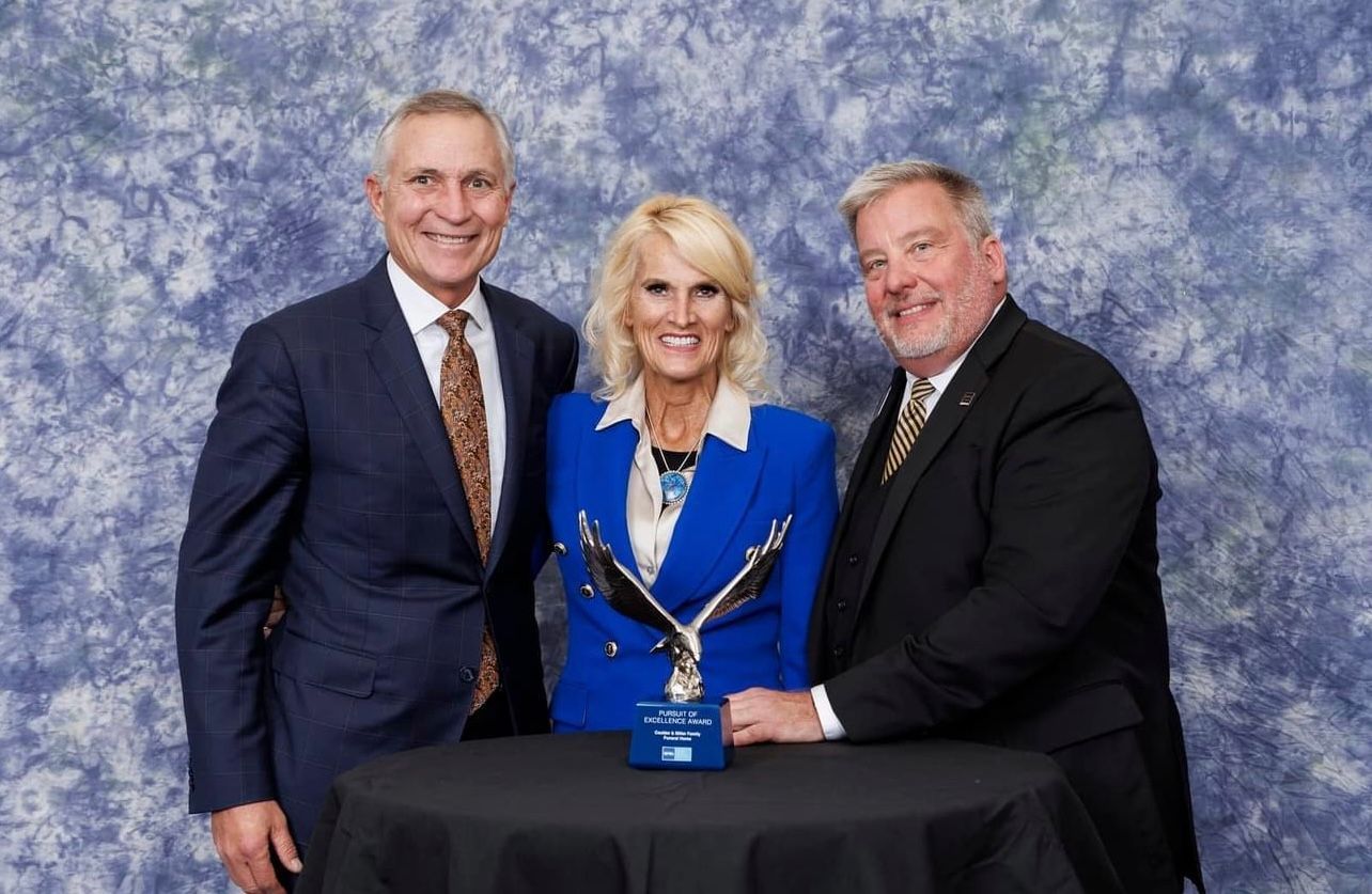 Three people posing with a blue award on a black table against a mottled blue backdrop.