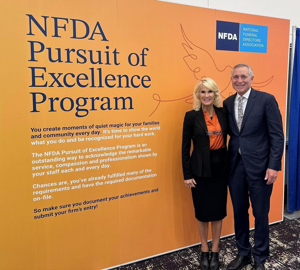 Two people standing by an orange NFDA Pursuit of Excellence Program display wall