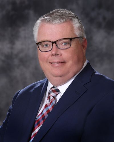 Portrait of a man in a navy suit, striped tie, and glasses against a gray backdrop
