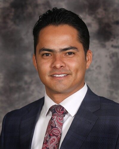 Portrait of a man in a dark suit, white shirt, and patterned tie against a gray studio backdrop
