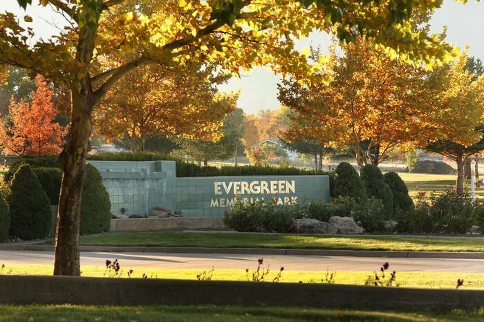 Evergreen memorial park entrance with autumn trees, stone sign, and sunlit lawn.