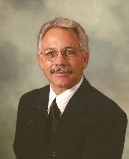 Studio portrait of a man in a black suit, white shirt, and tie, wearing glasses against a soft gray background