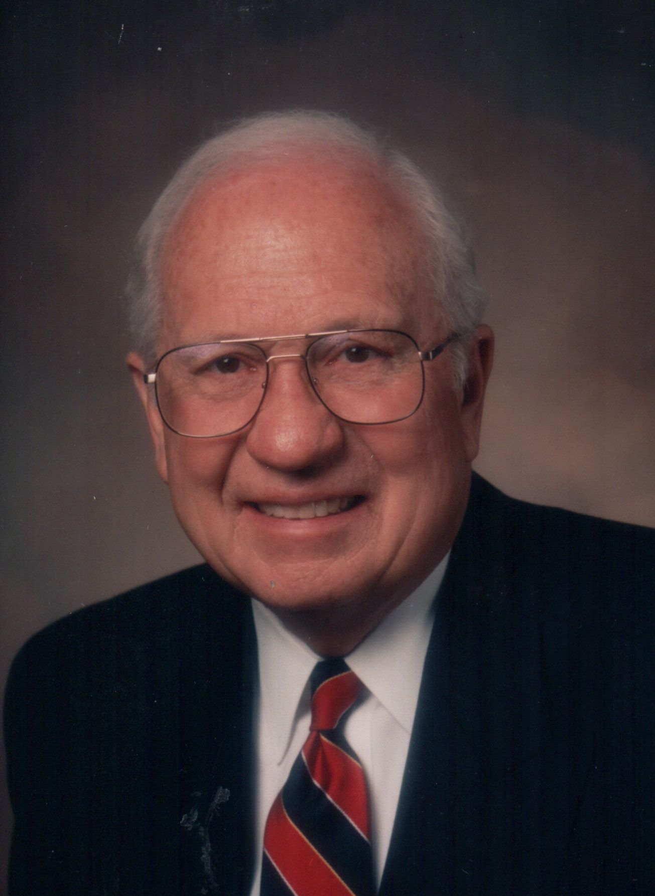 Smiling man in a dark suit, striped red tie, and glasses against a neutral studio backdrop