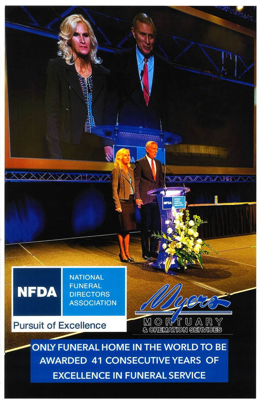 Award ceremony on a stage with two speakers, blue backdrop, flowers, and an award sign for NFDA/Hatch's Funeral Home.