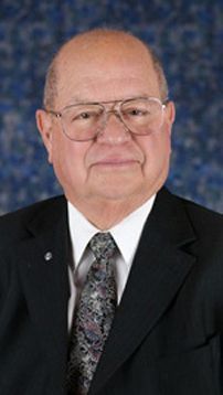 Formal portrait of a man in a dark suit and patterned tie against a blue backdrop
