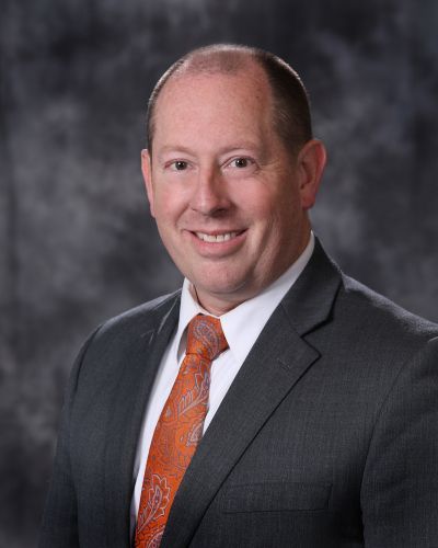 Studio portrait of a smiling man in a dark suit and orange tie against a gray background