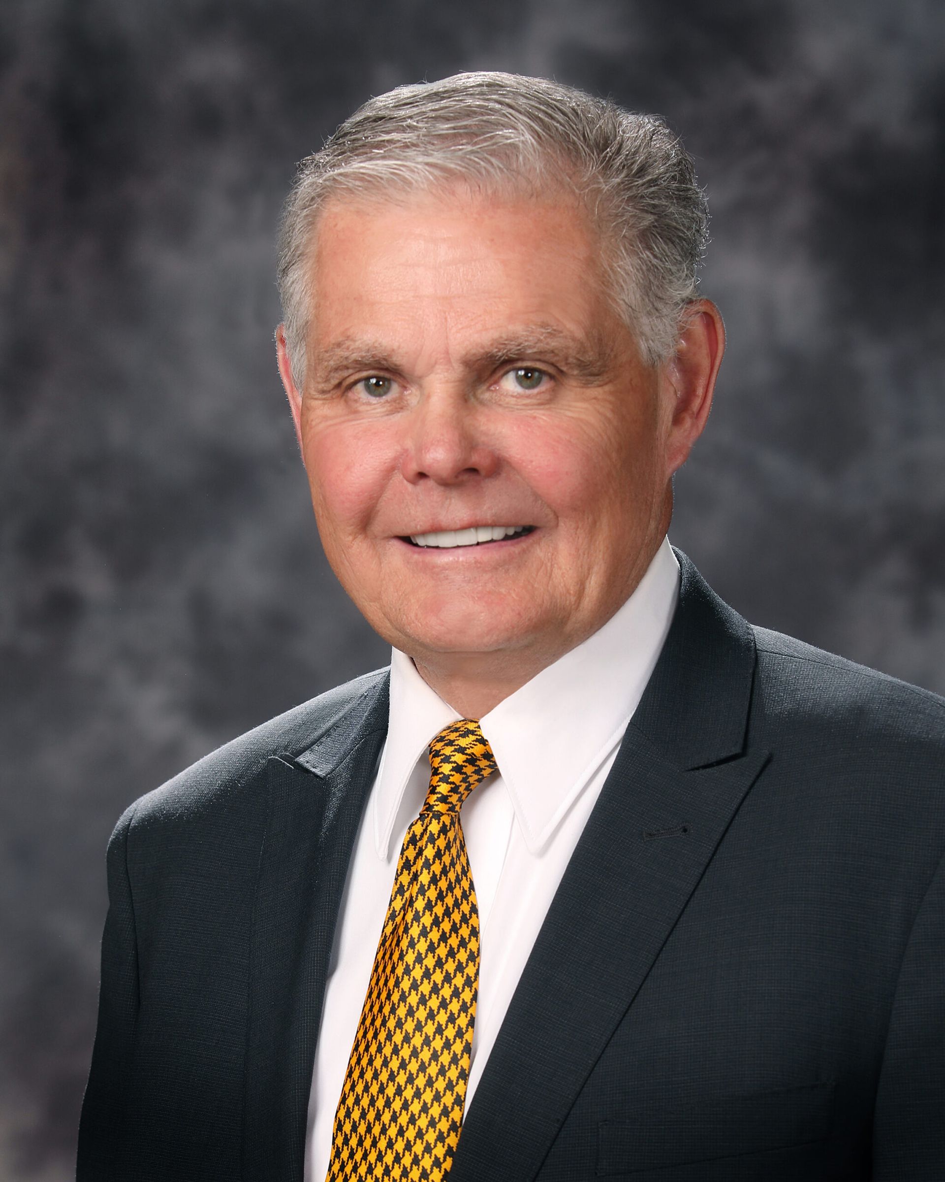 Studio headshot of a smiling man in a dark suit, white shirt, and gold tie against a gray backdrop