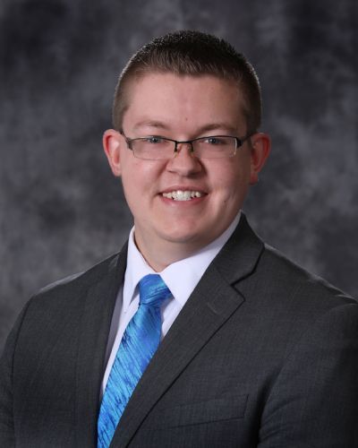 Studio portrait of a smiling person in a dark suit, blue tie, and glasses against a gray backdrop