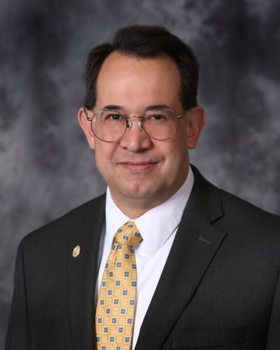 Portrait of a man in a dark suit, white shirt, and yellow tie against a gray backdrop