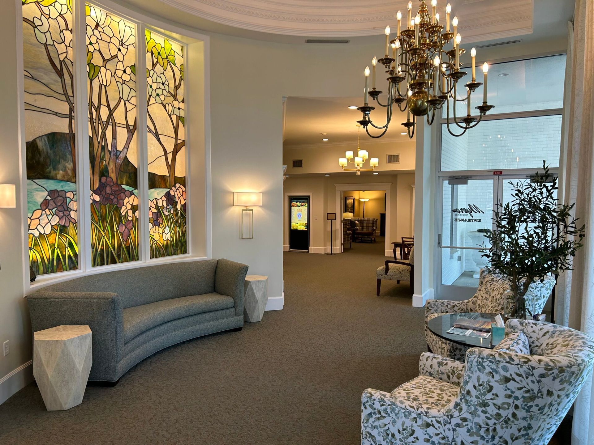 Elegant lobby with curved gray sofa, chandelier, floral chairs, and stained-glass windows