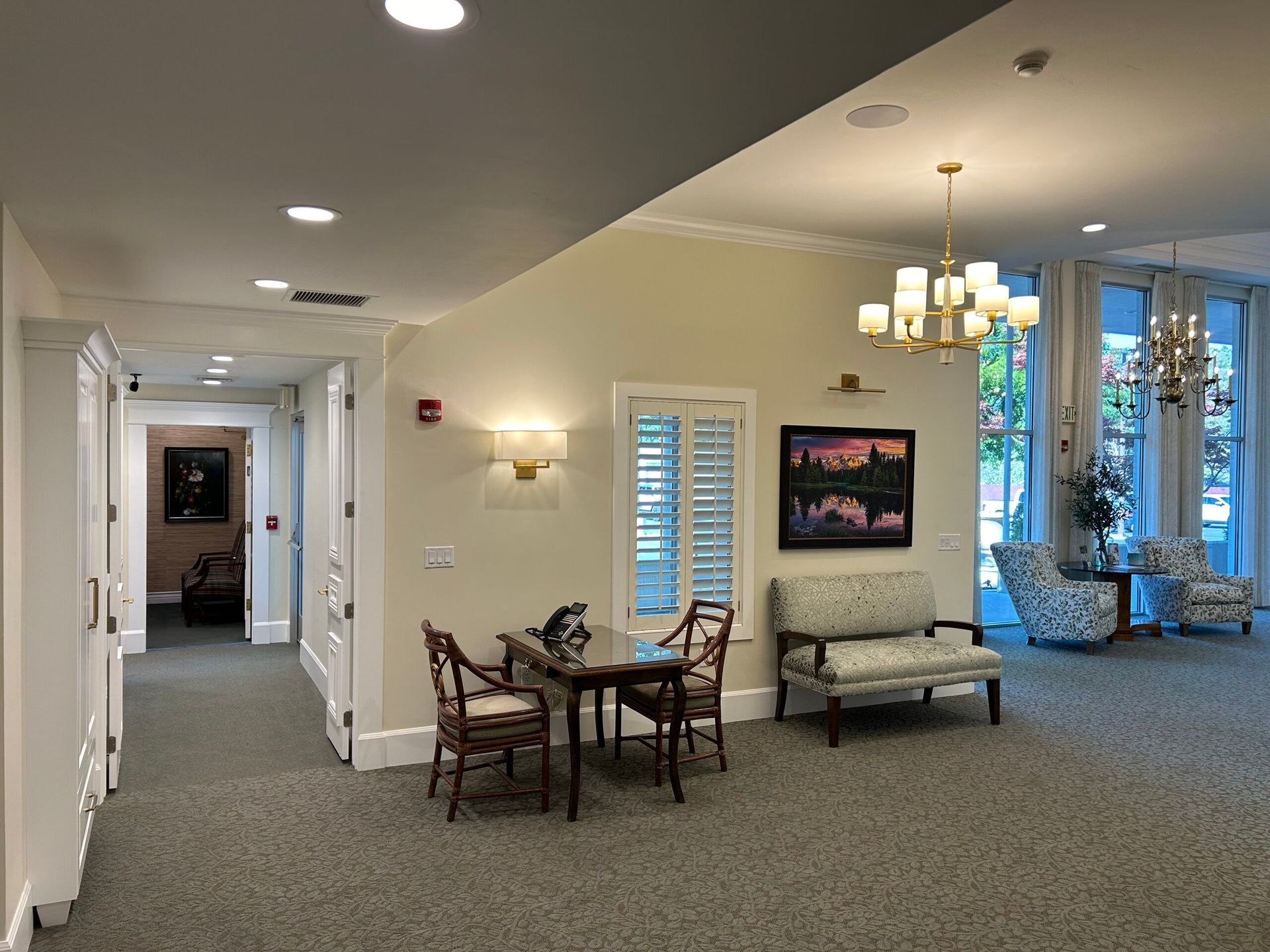 Bright hallway lounge with chairs, sofa, wall art, and a lit chandelier in a carpeted building interior