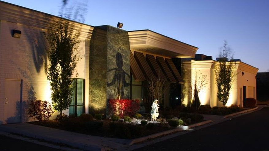 Night-lit building entrance with stone facade, arched canopy, and landscaped shrubs along the walkway