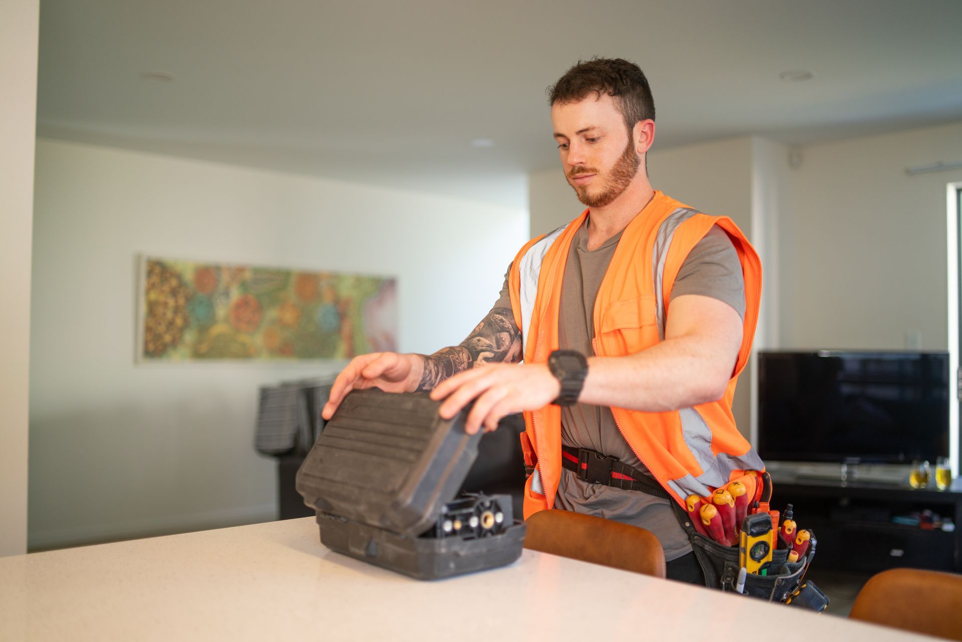 A man in an orange vest is sitting at a table holding a toolbox.