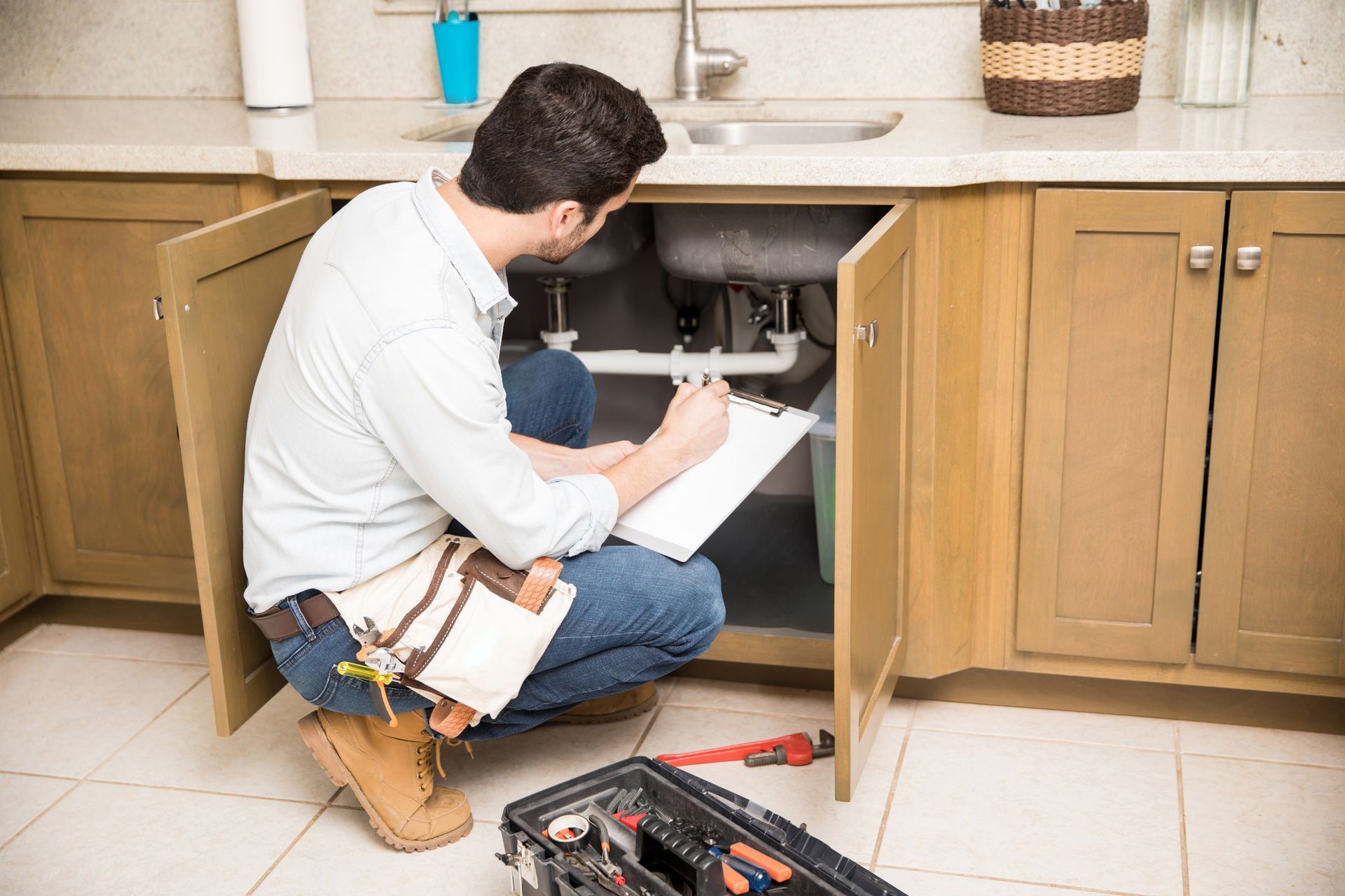 A man is kneeling under a sink in a kitchen and writing on a clipboard.