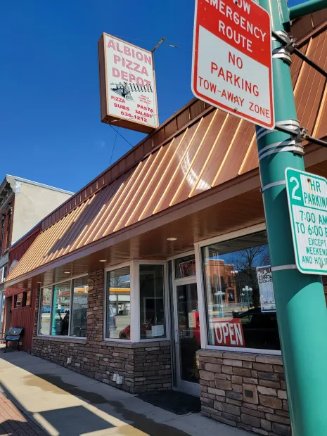 Storefront with striped awning, stone facade, and red signs for pizza, emergency route, and no parking.
