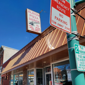 Storefront with striped awning, stone facade, and red signs for pizza, emergency route, and no parking.