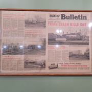 Framed newspaper bulletin on a pale green wall, with photos and red headline text.