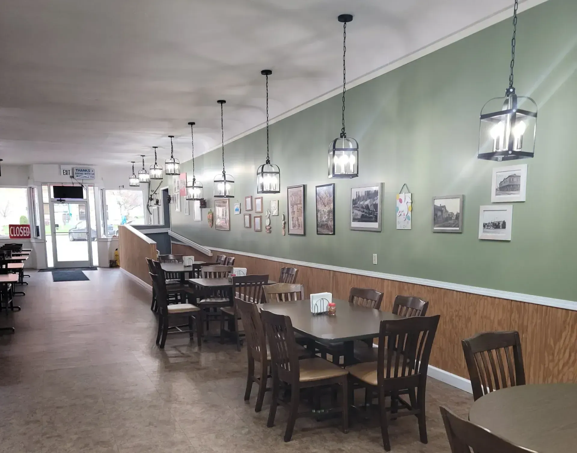 Empty restaurant dining room with long tables, hanging lights, and framed wall art along a green wall.