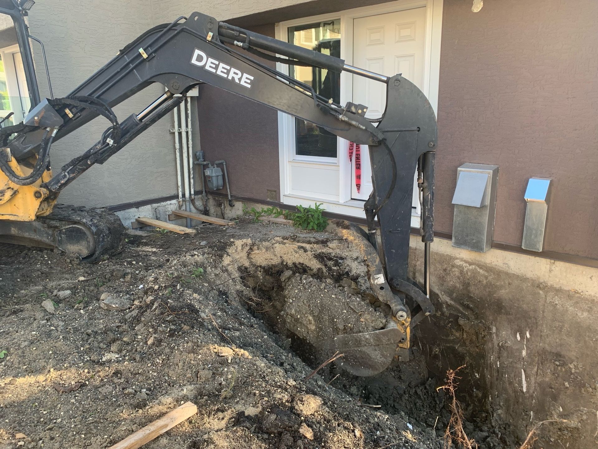 John Deere backhoe digging near a building's foundation, earth, and some green plants visible.