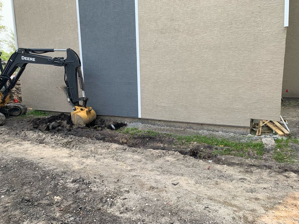 Small excavator digging a trench along a building exterior with beige and gray siding.