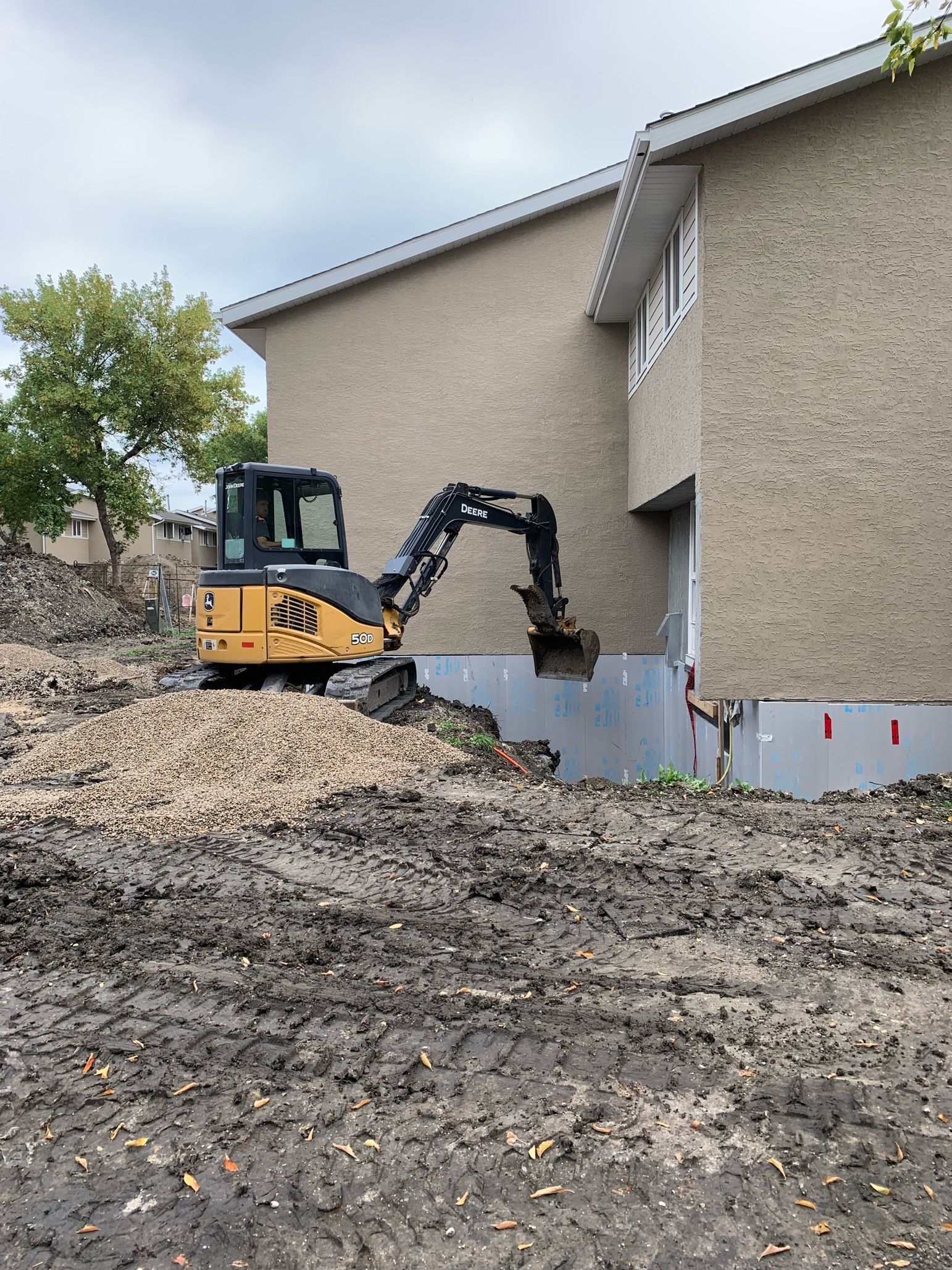 Mini excavator digging dirt near a house with beige stucco siding under a cloudy sky.