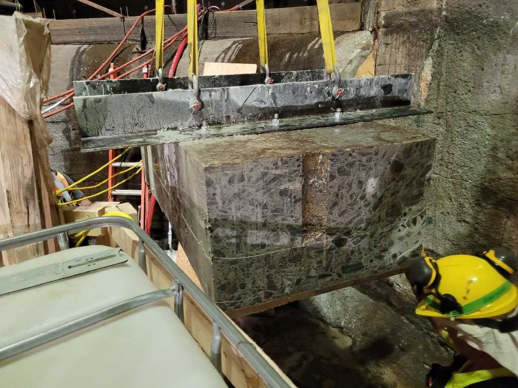 A large, concrete block suspended by yellow straps and a metal frame; setting appears industrial.