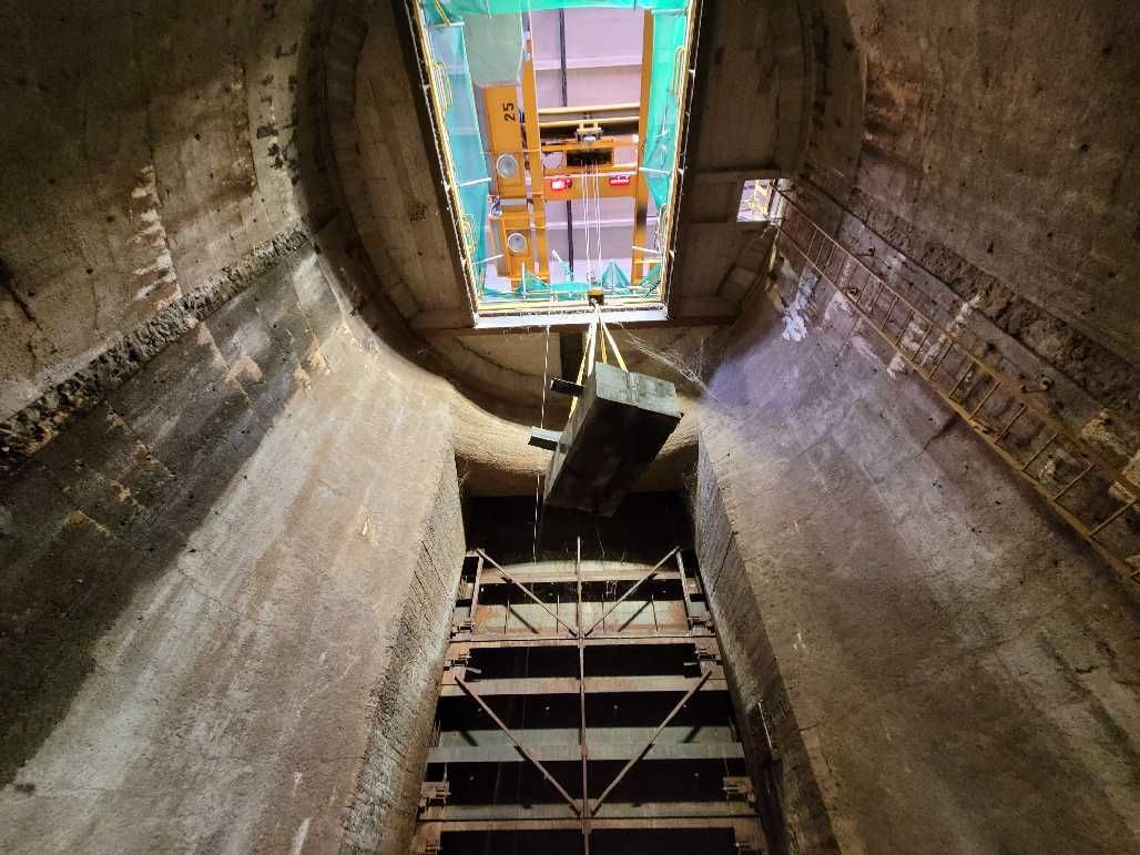 Inside view of a large, cylindrical, concrete tunnel with a ladder structure leading upwards toward a square opening covered by a screen.