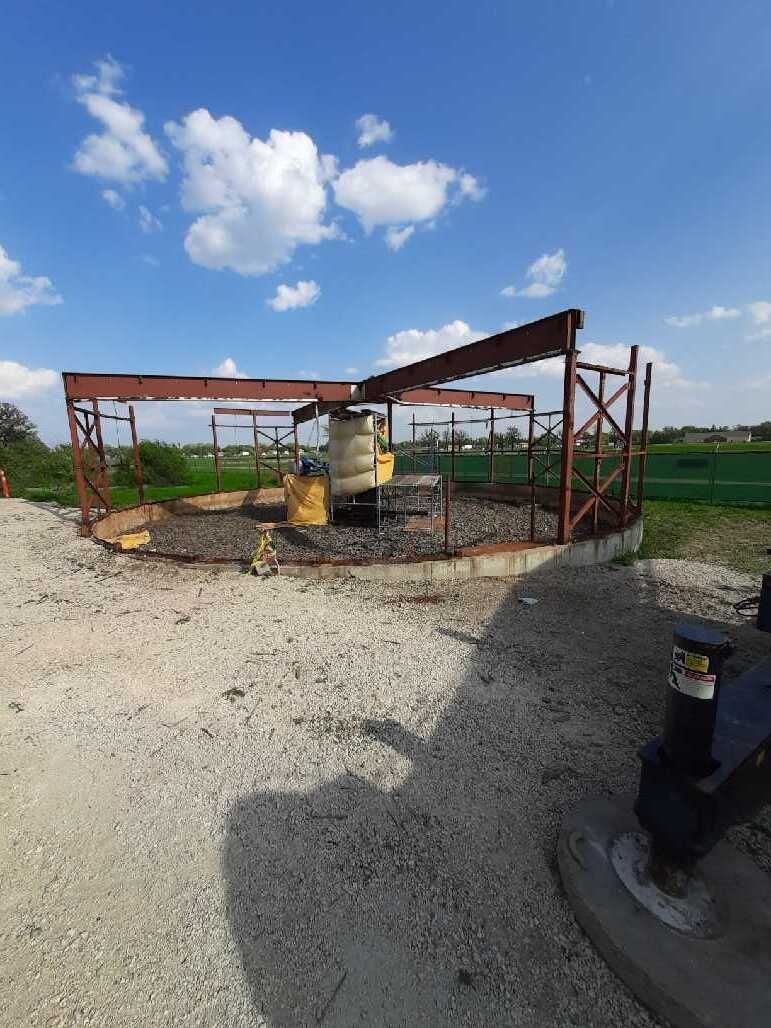 Construction site: metal framework surrounding cylindrical structure, gravel ground, blue sky with clouds.