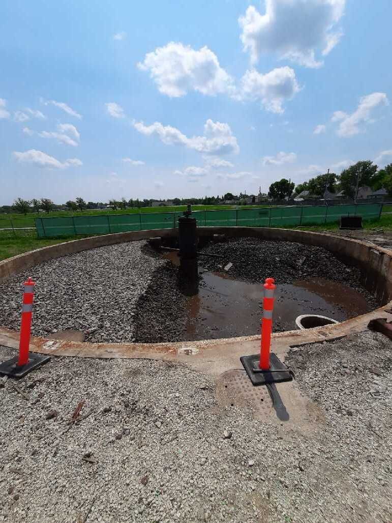 Circular wastewater treatment structure under construction with gravel and orange cones, set in an open field under a blue sky.