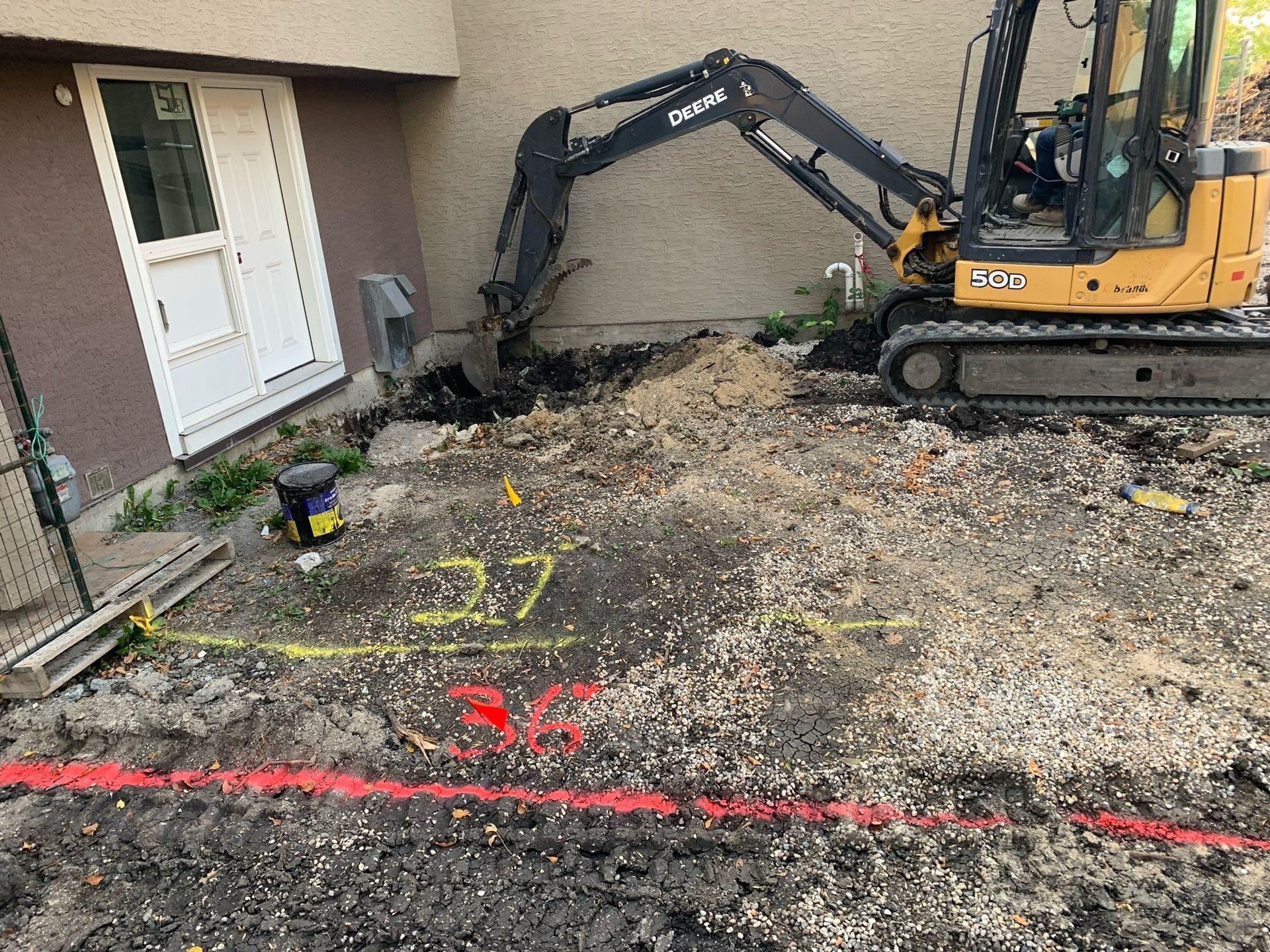 Mini-excavator digging near a building's white door. Marked ground with red and yellow lines, construction site.