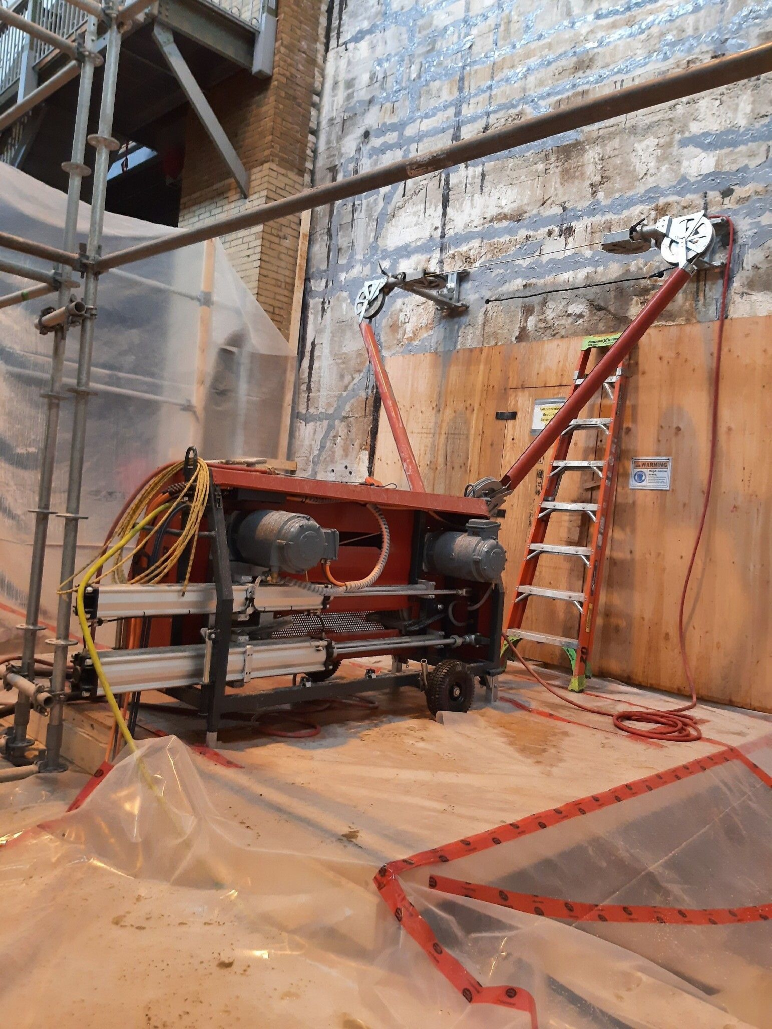 Red concrete saw cutting a wall, with scaffolding, ladder, and plastic sheeting in construction site.