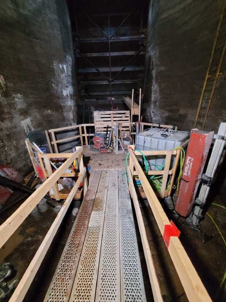 Narrow tunnel with metal walkway and equipment; wooden handrails line the path.