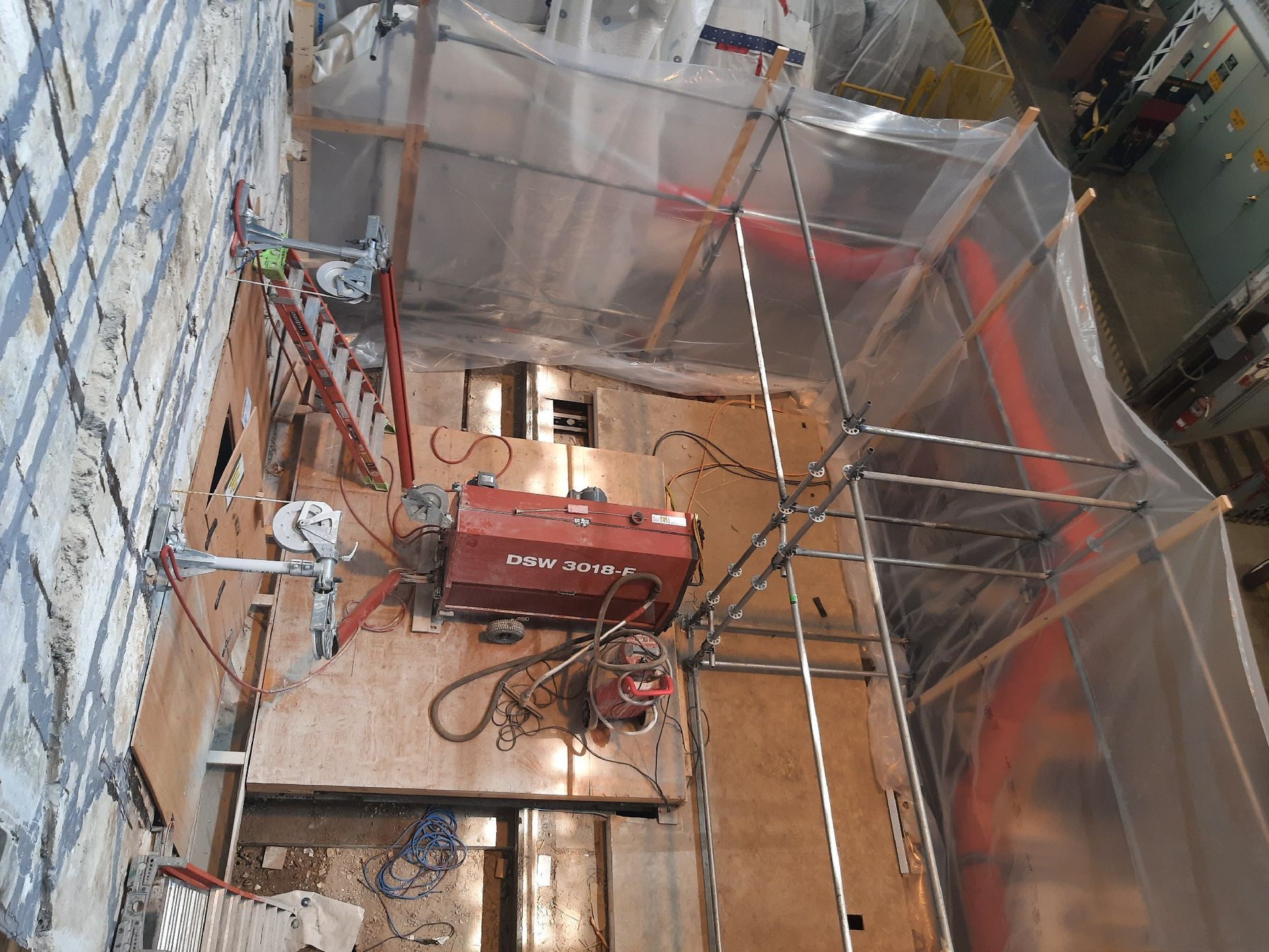 Construction site: Red saw equipment mounted on wall, scaffolding, plastic sheeting overhead.