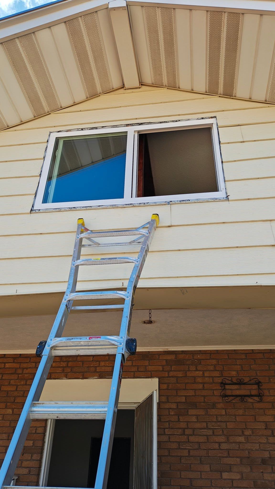 Blue ladder leaning against a house beneath an open window on a beige-sided exterior wall.