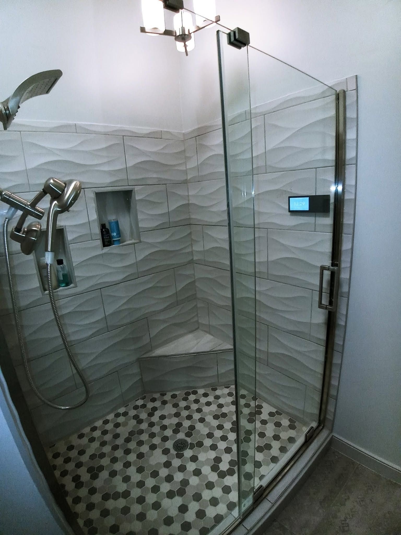 Modern glass shower stall with gray wavy tile walls, mosaic floor, handheld shower, and wall controls
