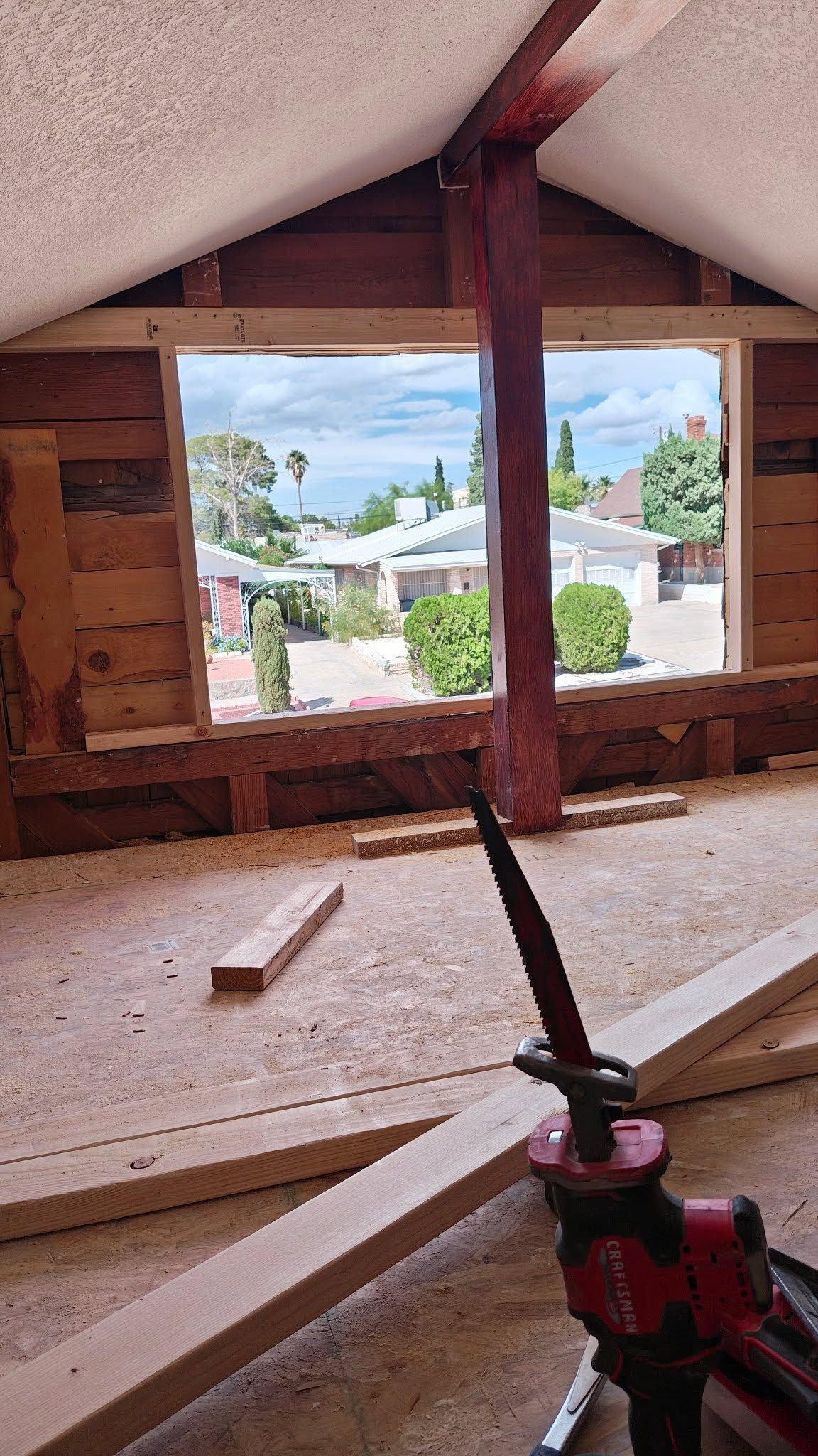 Attic under construction with exposed wood framing, open window, and a red power tool in the foreground