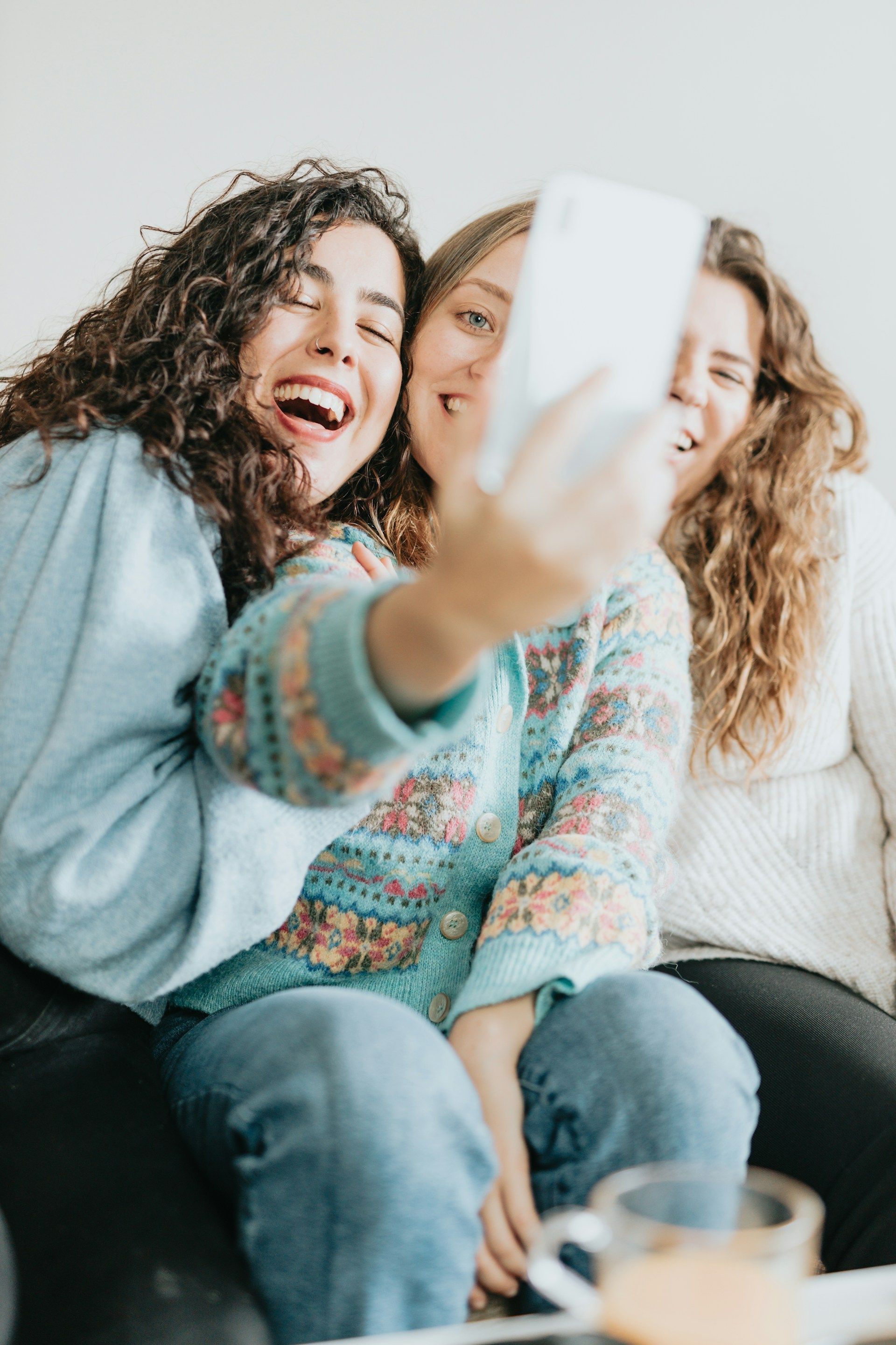 Three smiling friends take a selfie; one with curly hair laughs, the others smile; indoors.