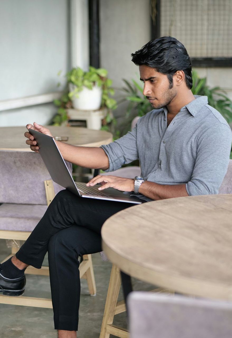 Man sits at a cafe table, typing on a laptop. Wearing a gray shirt and black pants.