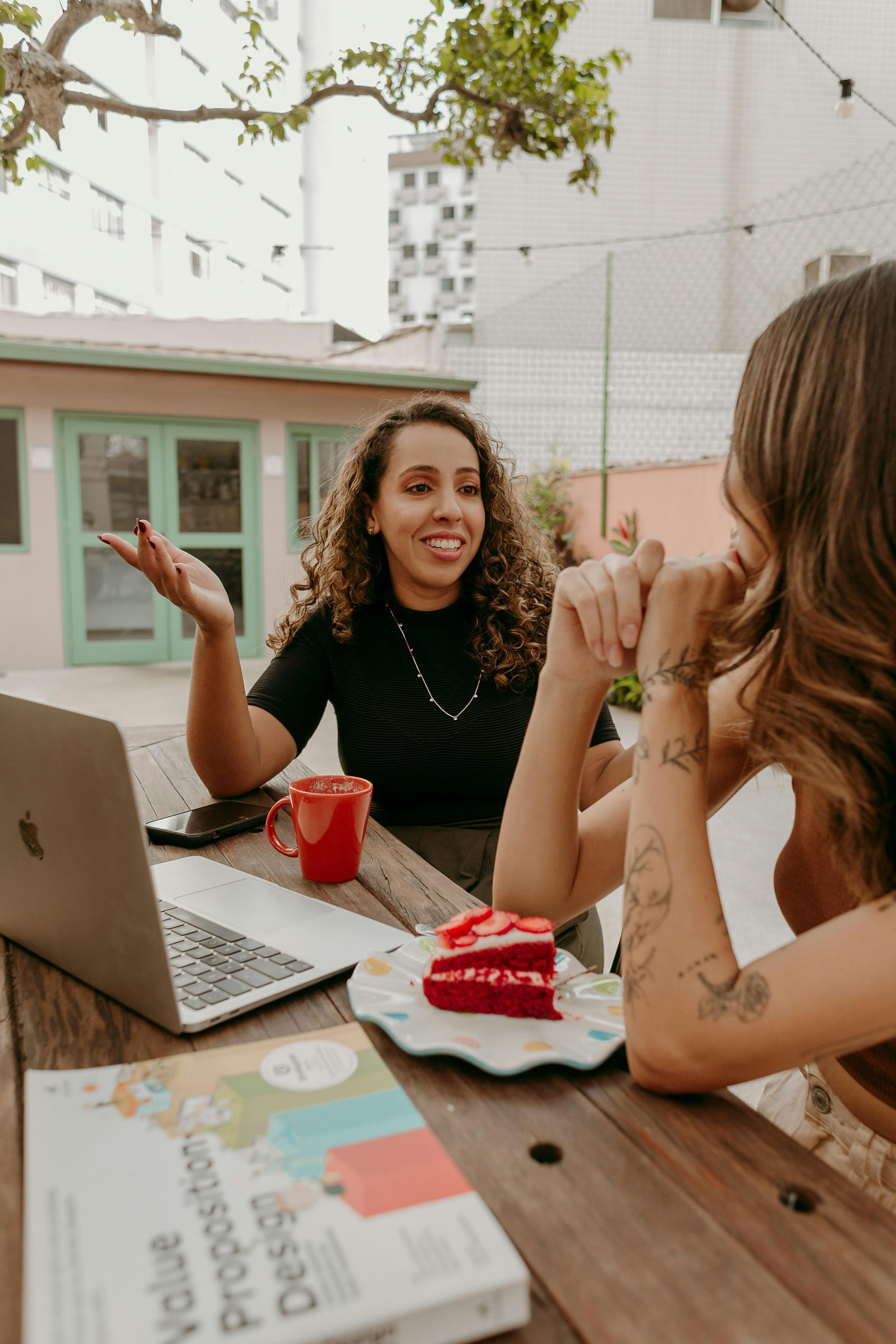 Two women at a table with laptop, book, coffee, and cake; one woman gestures and smiles. Outdoor setting.