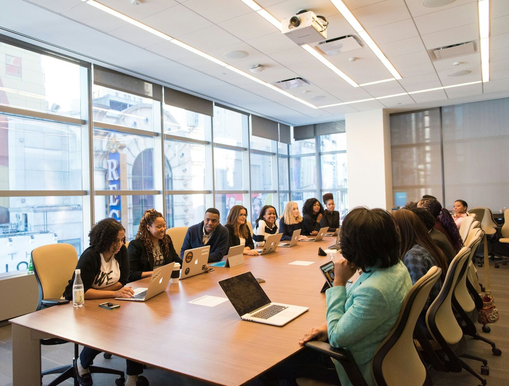 People at a meeting in a conference room with laptops, windows, and a projector.