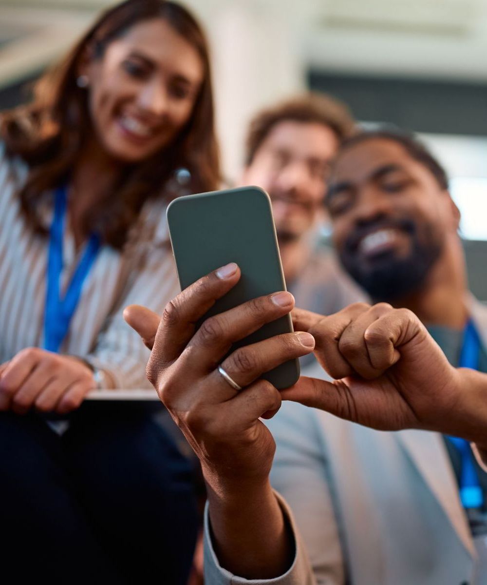 Three people smiling and looking at a smartphone held by a person with a ring on their finger.