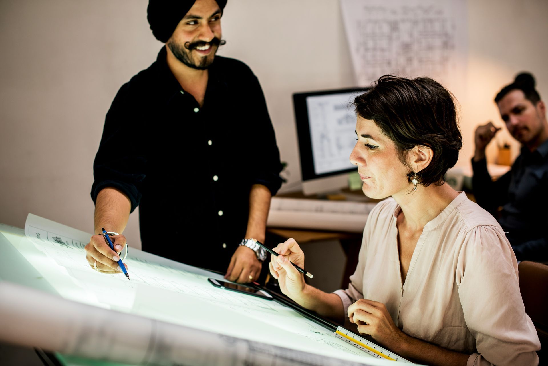 Two people working on a project at a light table, one pointing, the other looking on, another person in the background.