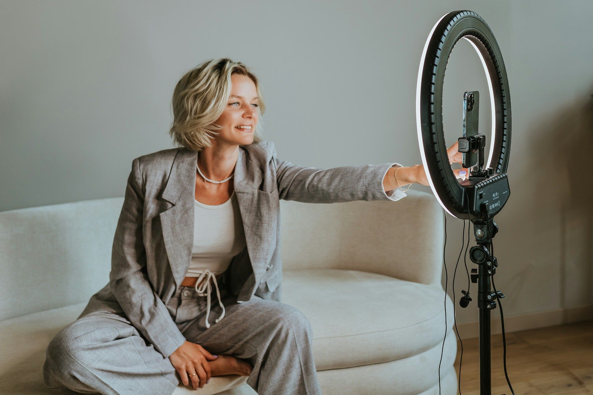 Woman seated on sofa, adjusting ring light, smiling. Wearing gray suit, white top, inside a room.