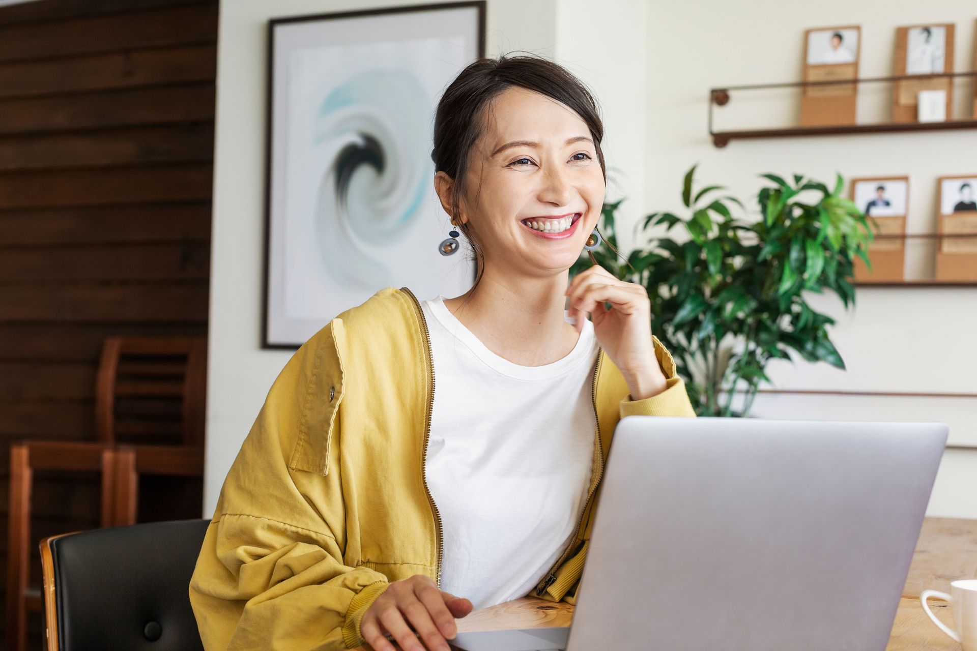 Woman smiles, working on laptop at a table, yellow cardigan, bright office setting.