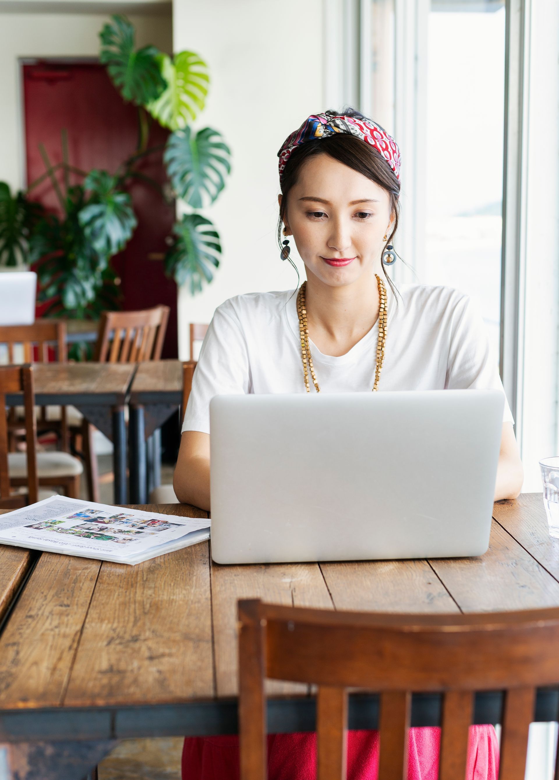 Woman using a laptop at a wooden table in a cafe, looking down. Floral headband, gold necklace, plants visible.