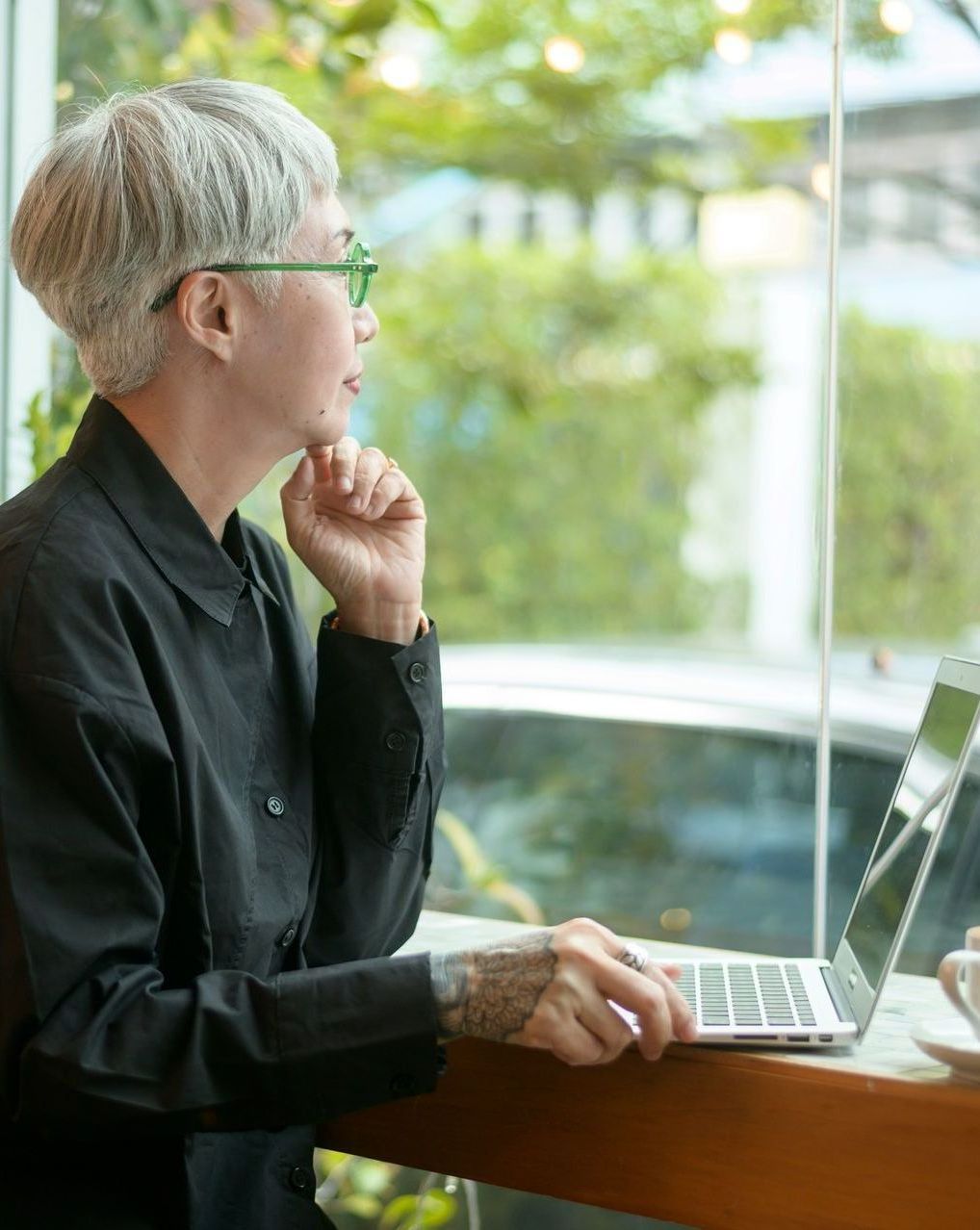 Person with grey hair and glasses looks out window, hand on chin, using laptop at table.