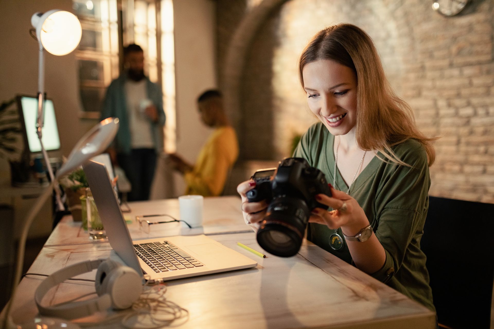Woman examines a camera, smiling, at a desk with a laptop and headphones. Two people are in the background.