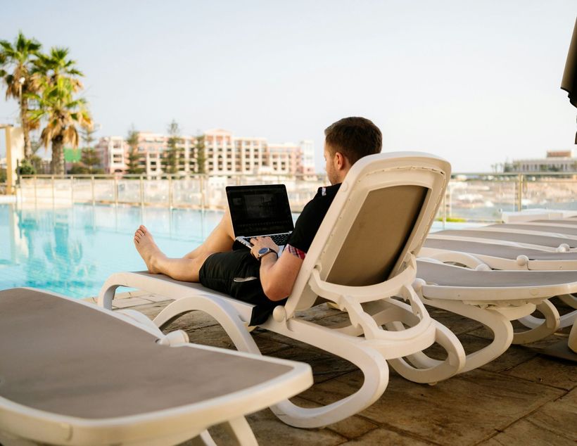 Man working on a laptop while relaxing on a lounge chair by a pool, with palm trees and a building in the background.