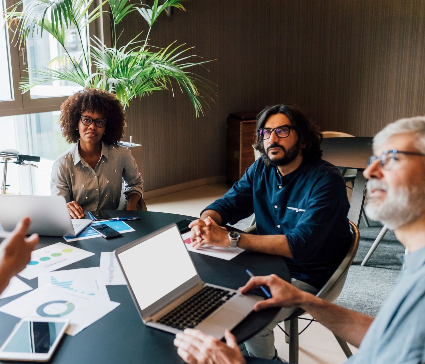 Group of people in meeting around a table with laptops, charts and a plant, indoors.
